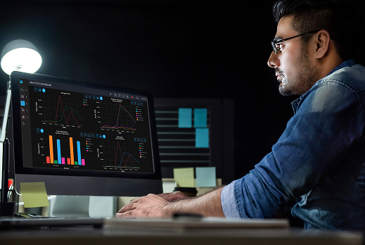 Person working at a desk with a computer monitor displaying multiple charts and graphs for data analytics, including bar charts, line graphs, and performance metrics in a dark-themed interface.