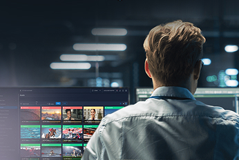 Person seated at a workstation in a broadcast or media control room, viewing multiple monitors displaying video thumbnails of sports, news, and entertainment content on a large screen interface