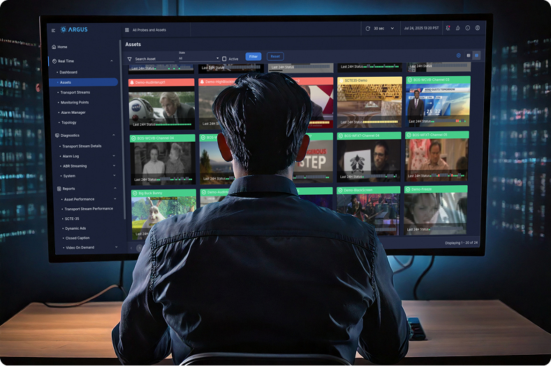 Person sitting at a desk in front of a large monitor displaying a media asset management interface with multiple video thumbnails and navigation options