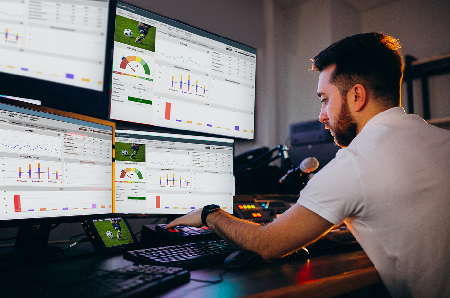 Person working at a desk with multiple monitors displaying sports analytics dashboards, including charts, graphs, and performance metrics for soccer matches