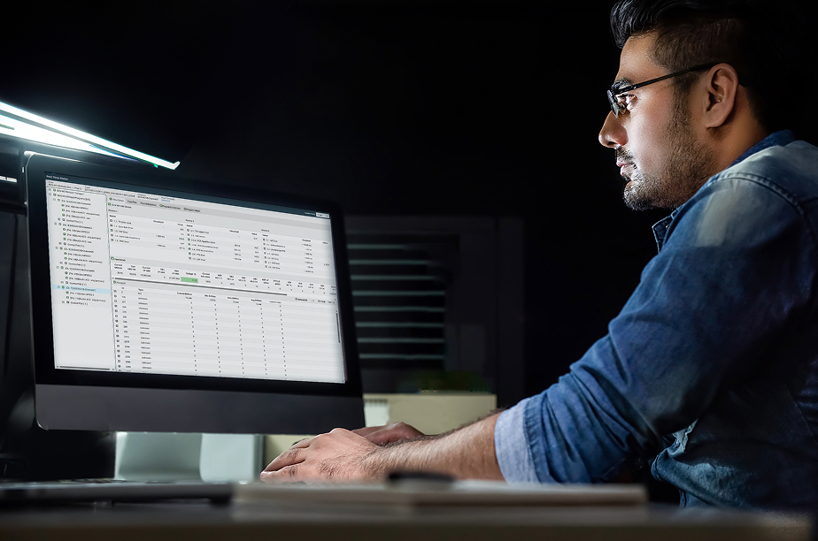 Person working at a desk with a computer monitor displaying a data management interface with tables, columns, and numerical values in a dark office setting