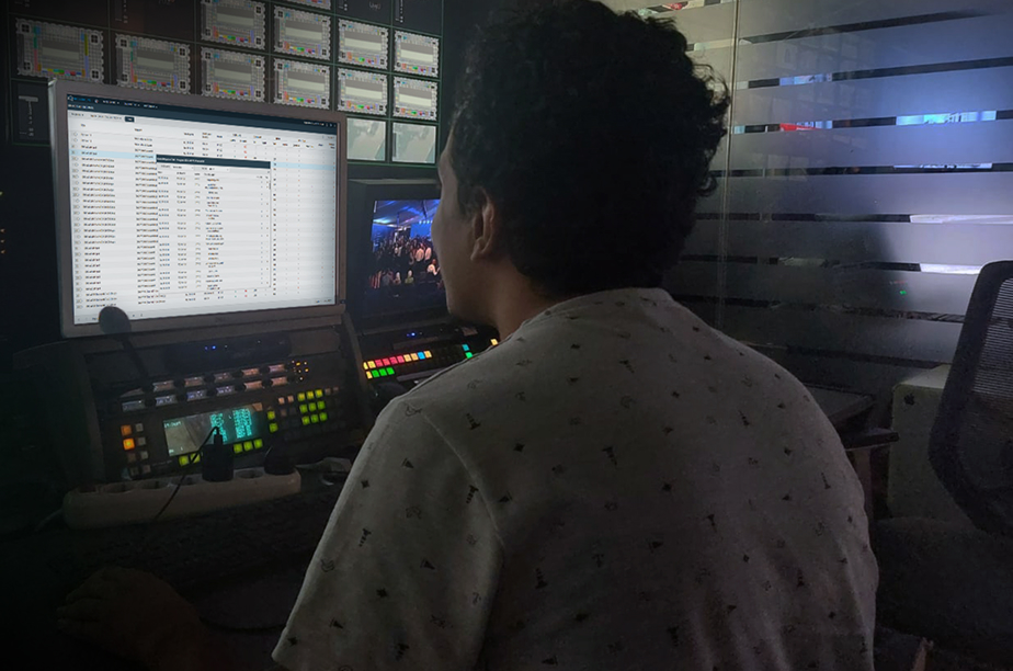 Person working at a broadcast control station with a computer monitor displaying a compliance and subtitle management interface, alongside a smaller screen showing a live event feed