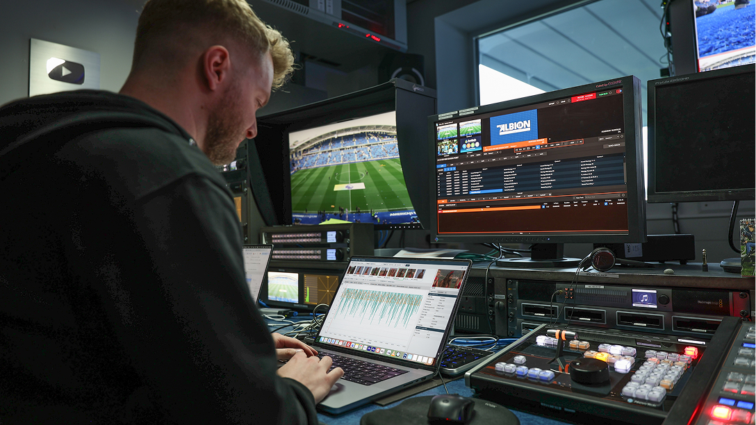 Broadcast control room setup with multiple monitors displaying a soccer stadium feed and video editing software, alongside a laptop showing audio waveform analysis and a professional video switcher