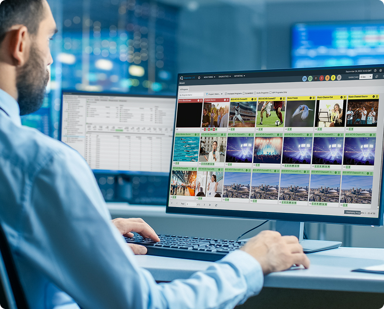 Person working at a desk with a computer monitor displaying a media monitoring interface featuring multiple video thumbnails of sports, entertainment, and news content, alongside a secondary screen showing data tables
