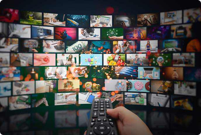 Hand holding a remote control pointed at a large television screen displaying a grid of colorful thumbnails from various streaming services and entertainment options.