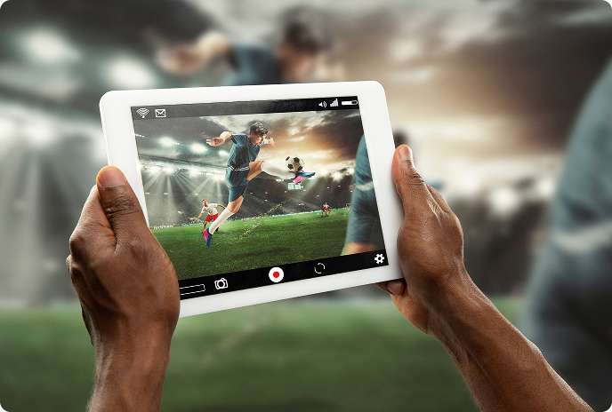 Hands holding a tablet streaming a live soccer match, showing a player kicking a ball on a brightly lit stadium field