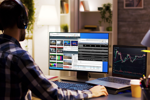 Person working at a desk with a large monitor displaying multiple video thumbnails and signal analysis tools, alongside a laptop showing a financial chart, in a modern home office setting.