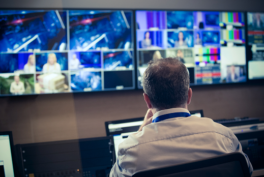 Broadcast control room with a person seated at a workstation monitoring multiple large screens displaying live video feeds, color bars, and production elements for television broadcasting.