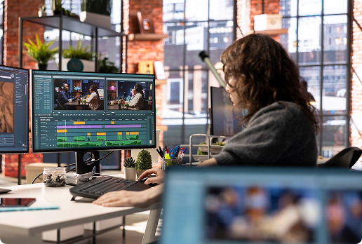 Person working at a desk in a modern office environment using a computer with dual monitors displaying video editing software and timeline tracks.