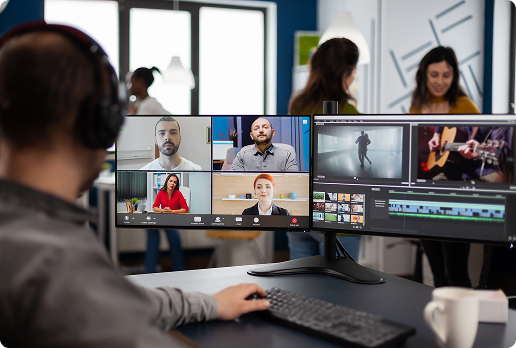 Person wearing headphones working at a desk with dual monitors showing a video conferencing interface on one screen and video editing software on the other, in a modern office environment.