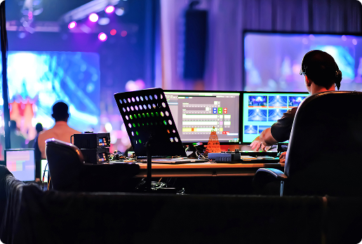 Live event production setup with a person operating multiple monitors and control panels, showing video feeds and switching interfaces in a stage environment with colorful lighting.