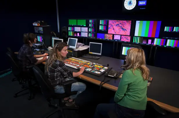 Broadcast control room with multiple monitors displaying color bars and video feeds, two operators seated at a large production switcher panel managing live video signals
