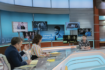 A television studio set with multiple screens displaying images and text, including one screen. Two people are seated at a curved desk with papers and notes in front of them, while another person sits across the desk being interviewed. The background features bright blue panels and several monitors with various images