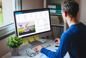 person sitting at a desk using a desktop computer, viewing a video editing or media management interface with a cartoon animation displayed on the screen. The desk has a keyboard, mouse, eyeglasses, sticky notes, and a potted plant, with shelves and binders in the background