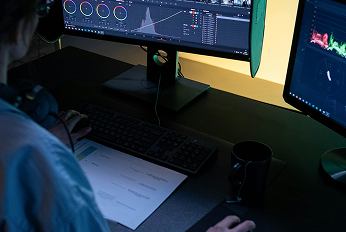 Person working at a desk with dual monitors displaying a video editing interface featuring color grading tools and waveform graphs, with a keyboard, paper document, and coffee mug on the desk.