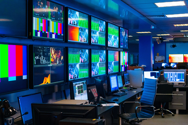 Broadcast control room with multiple monitors displaying live video feeds and color bars, featuring workstations with computers and control panels in a professional media production setting