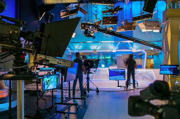 behind-the-scenes view of a television studio with cameras, monitors, and crew setting up under bright blue lighting