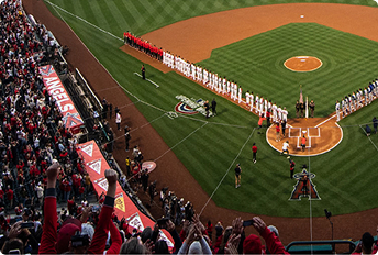Baseball stadium during a pre-game ceremony with players lined up along the first and third baselines, a color guard at home plate, and fans cheering in the stands.
