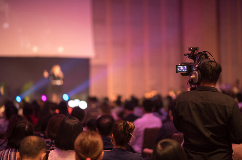 Videographer recording a live event in a large conference hall with an audience seated and a speaker on stage