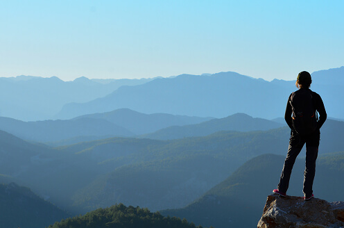 Person standing on a rocky peak overlooking a vast mountain range under a clear blue sky