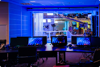 Broadcast control room with multiple monitors and equipment, overlooking a brightly lit television studio with cameras and lighting rigs