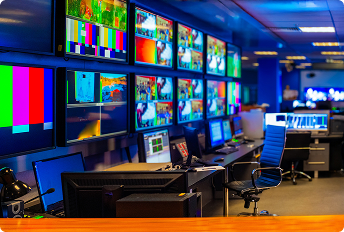 Broadcast control room with multiple large screens displaying color bars, live video feeds, and monitoring interfaces, alongside workstations equipped with computers and professional chairs.