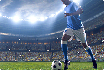 Soccer player in a blue jersey and white shorts dribbling a ball on a brightly lit stadium field with a large crowd in the background.