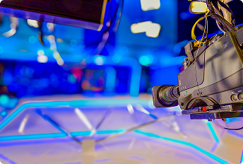 Close-up of a professional broadcast studio camera mounted on a rig, with blurred blue-lit studio background featuring modern lighting and equipment.