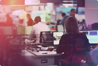 Modern newsroom environment with multiple people working at desks equipped with computers and monitors displaying video editing software and live broadcast content.