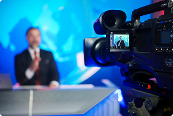 Professional video camera recording a news anchor at a studio desk with a blue-toned background and bright studio lighting