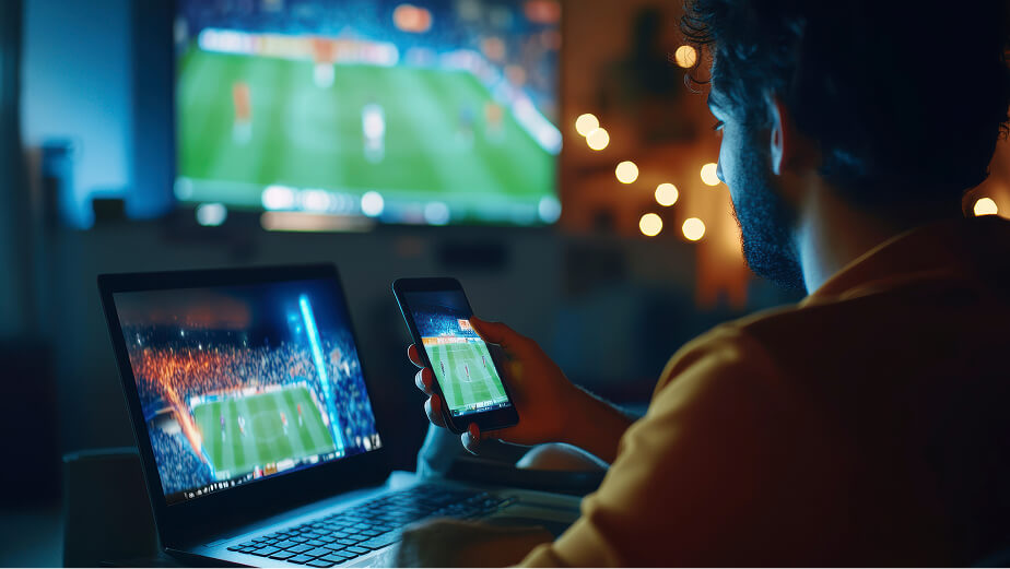 Person watching a soccer match on a large TV while simultaneously streaming the same game on a laptop and smartphone in a cozy, dimly lit room